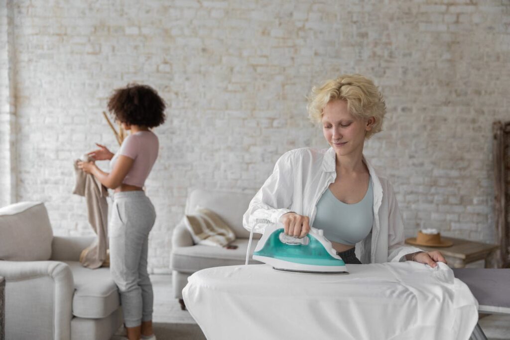 Busy woman ironing clothes and black girlfriend folding clothes during daily routine at home