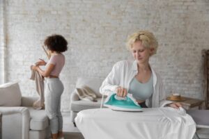 Busy woman ironing clothes and black girlfriend folding clothes during daily routine at home