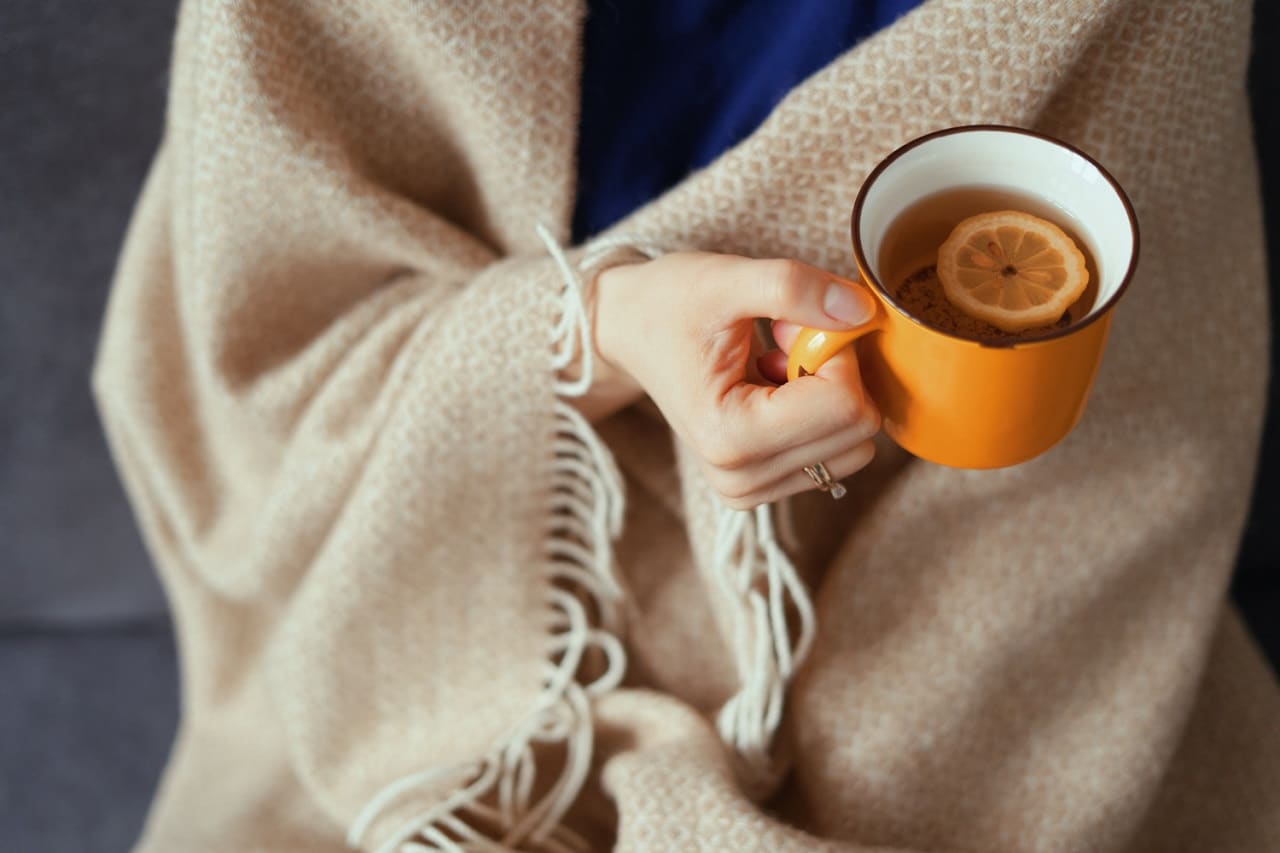 Une femme envelopper dans une couverture chauffante tenant une tasse de thé de couleur orange à la main