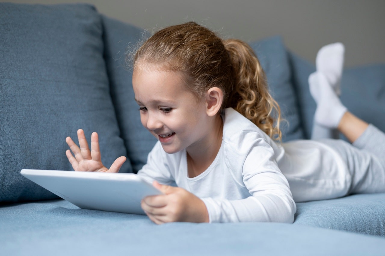 Une fillette blonde avec une queue de cheval utilise une tablette tactile allongée sur un canapé gris