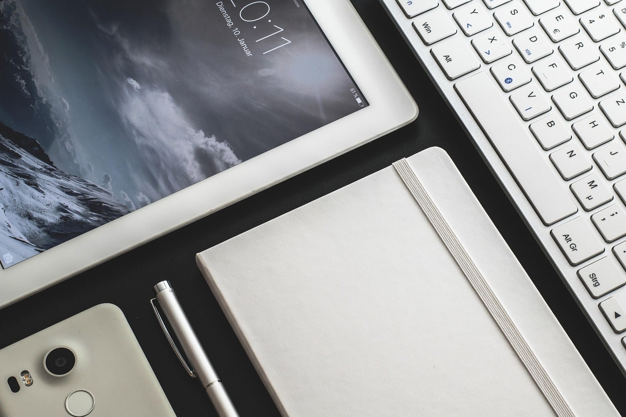 Sur l'image on observe une petite partie d'un bureau. On y voit une tablette électronique allumée sur l'écran de verrouillage, un carnet blanc et fermé, un stylo, un téléphone et un clavier d'ordinateur.