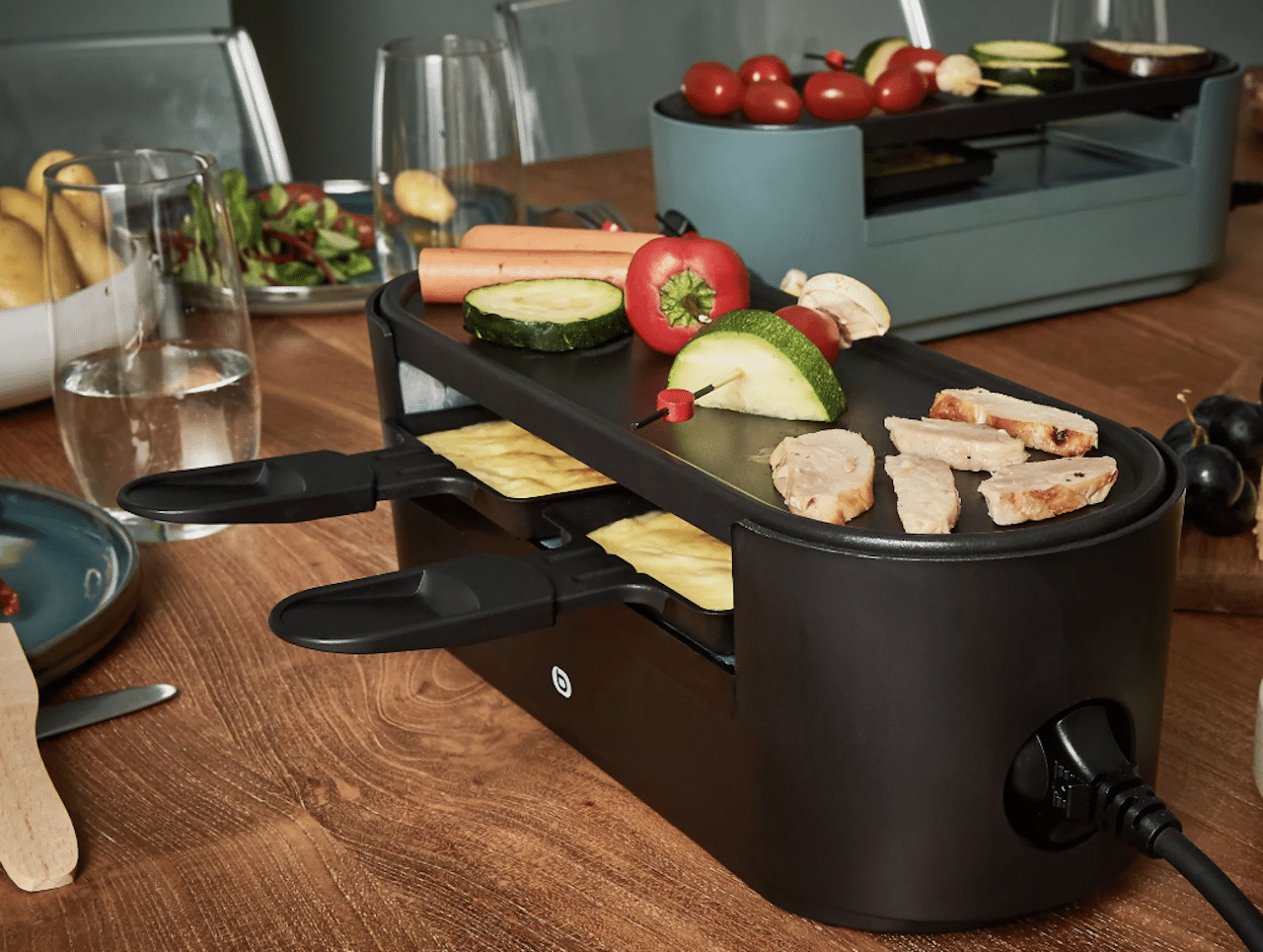 Un appareil à raclette de la marque Essentiel B posé sur une table à maner au milieu d'assiettes et de verres.
