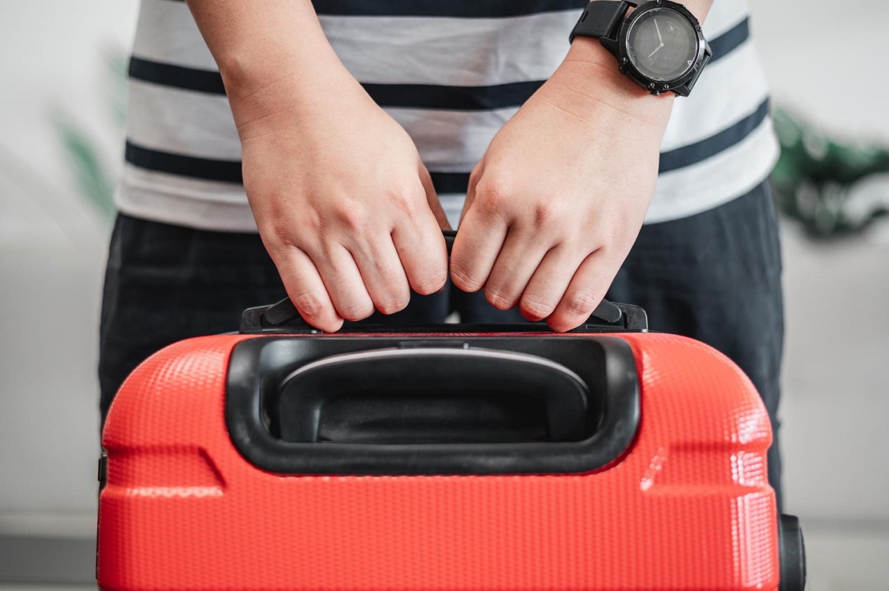 Une personne portant un T-shirt rayé soupèse une valise rouge avec ses 2 mains