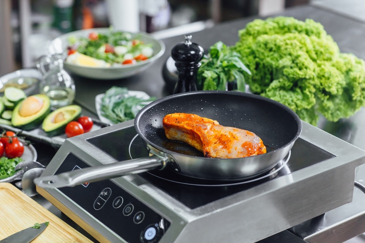 Un rechaud à induction posé sur un plan de travail avec une poele dans laquelle cuit une cote de porc