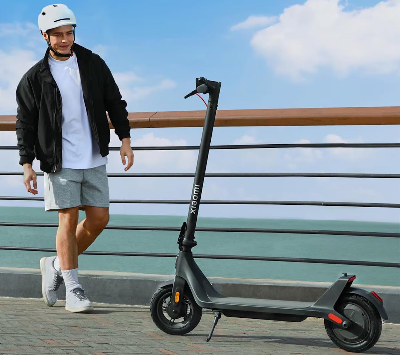 Un homme qui se tient à côté d'une trottinette électrique de la marque Xiaomi. Il est en extérieur près de la mer.