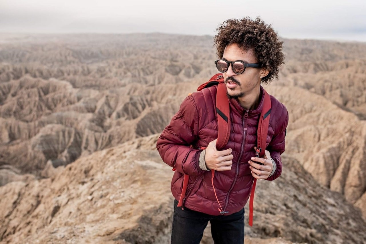Un randonneur avec un anorak rouge dans un paysage de montagne porte des lunettes Bose Frames Rondo