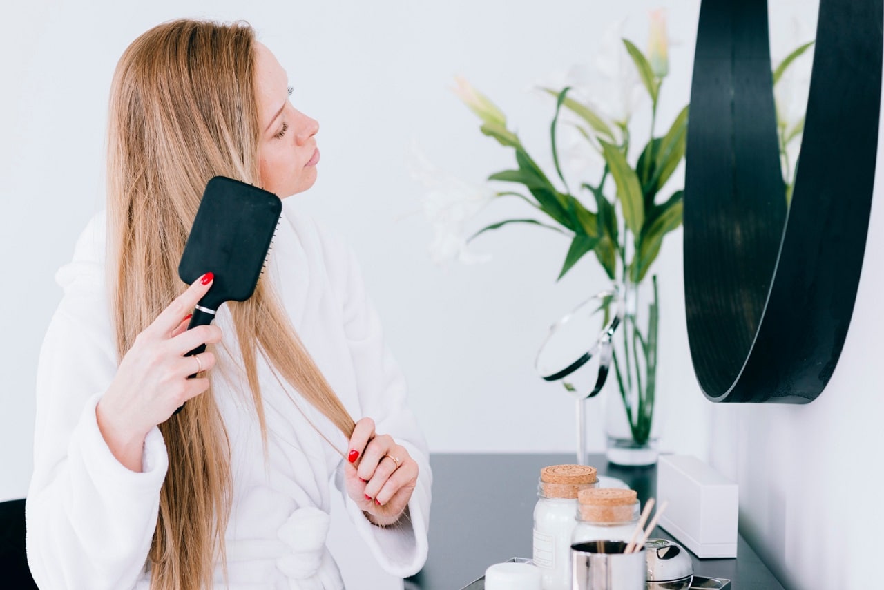 Une jeune femme blonde lisse ses cheveux longs avec une brosse lissante devant le miroir de sa salle de bain