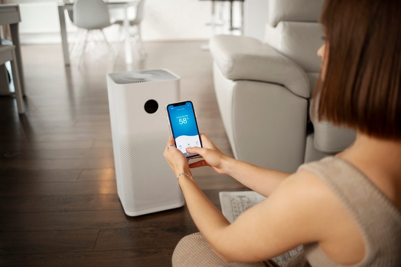 Une jeune femme assise sur son canapé règle son climatiseur mobile avec son téléphone mobile