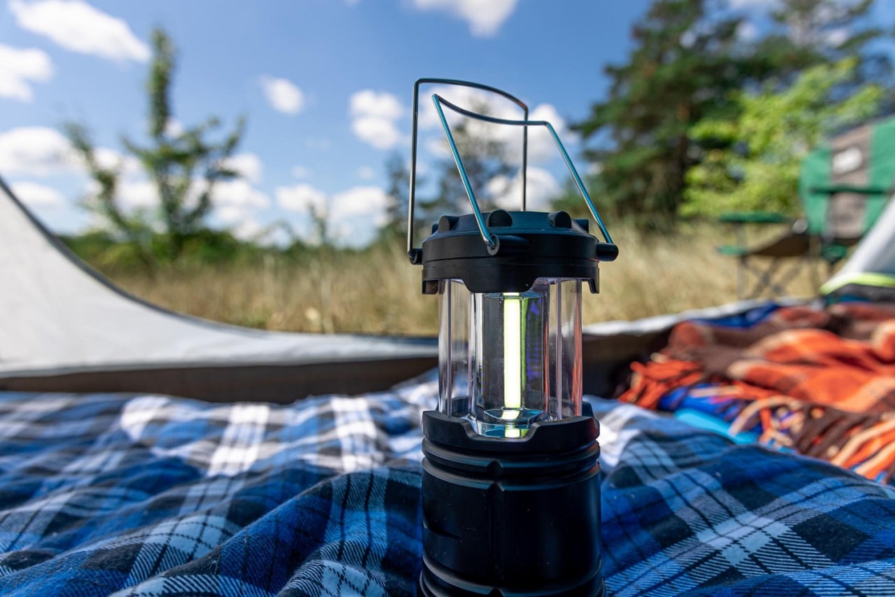Une lampe de camping posée sur un plaid dans une tente ouverte où l'on aperçoit le paysage de jour