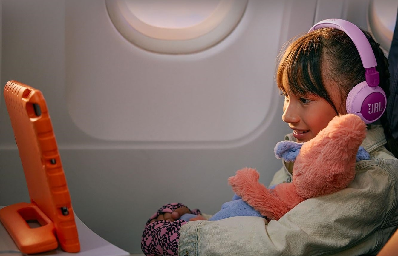 Une petite fille avec un ours en peluche rose dans les bras est assise dans un avion et regarde une video sur un écran portable avec un casque sans fil JBL sur les oreilles