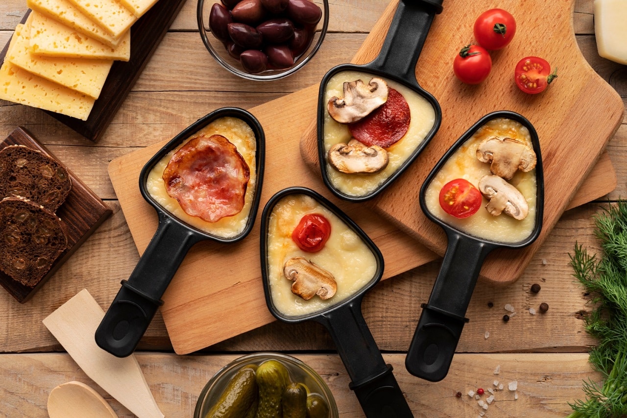4 coupelles à raclette contenant du fromage fondu recouvert de champignons, de bacon fumé, de chorizo et posées sur 2 planches en bois