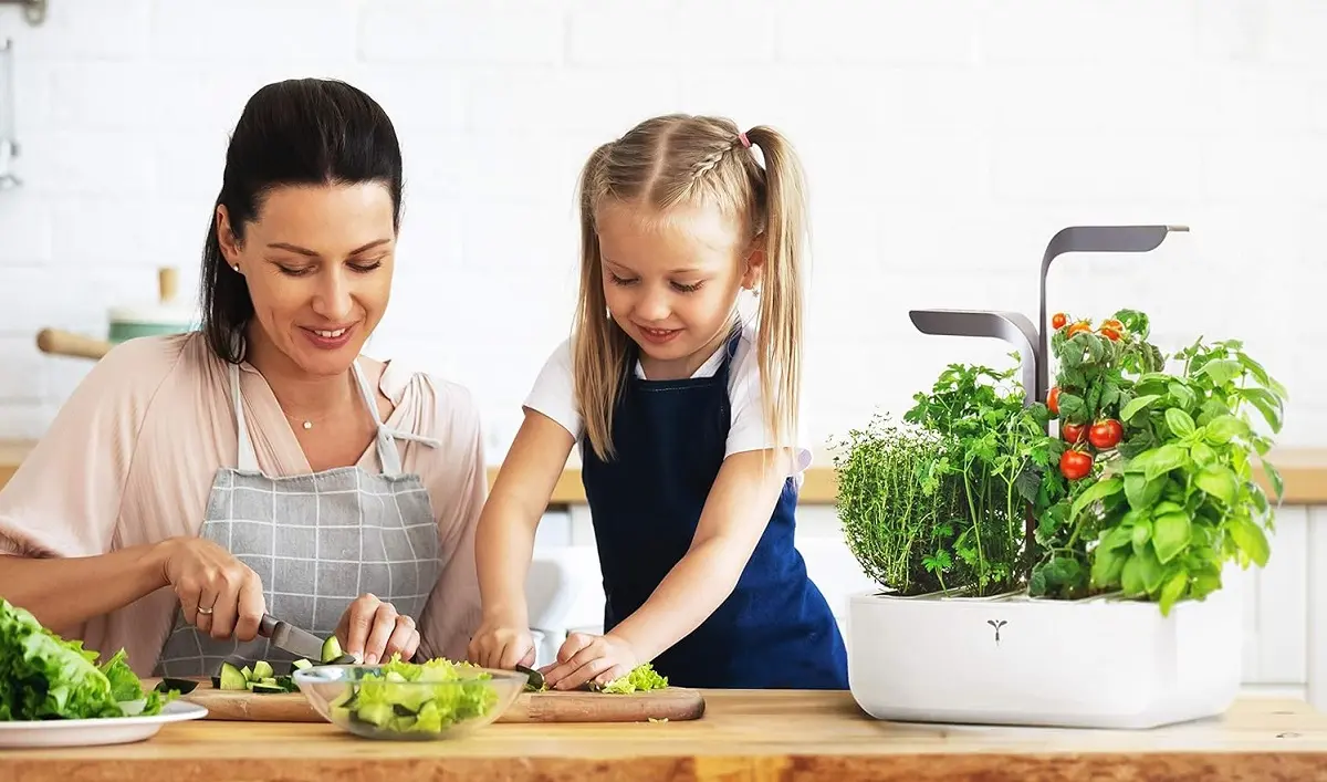 Une maman et sa petite fille coupe des plantes aromatiques sur un plan de travail sur lequel est posé le potager connecté Véritable.