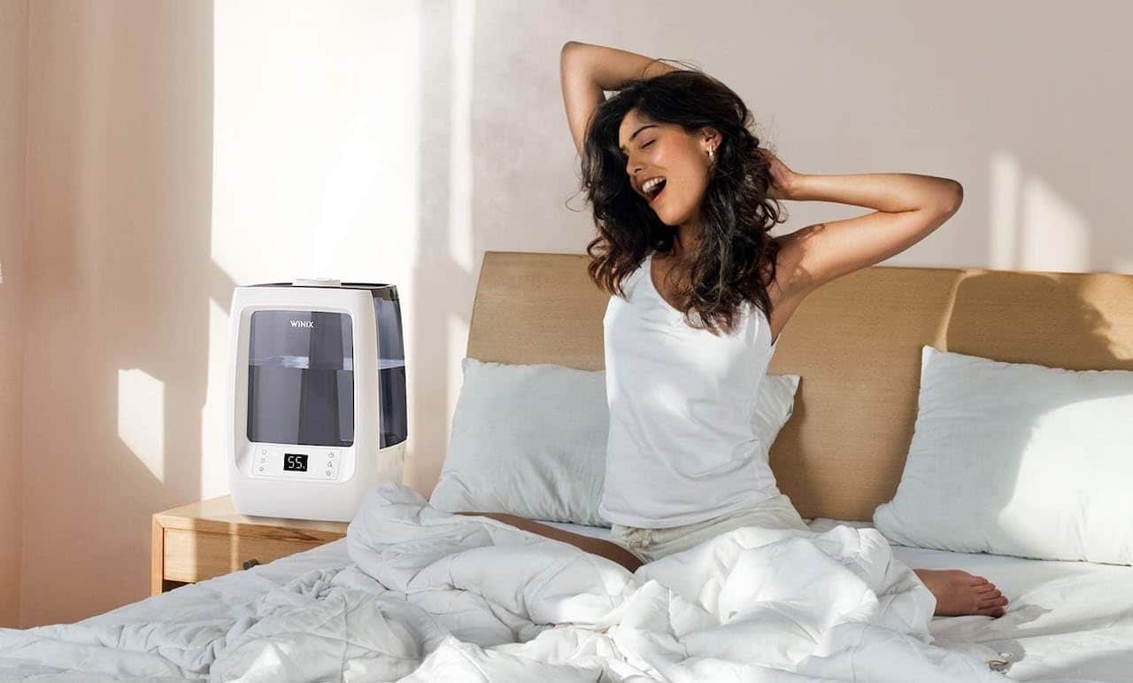 Une femme se réveille dans un lit double à côté d'un humidificateur d'air Winix.